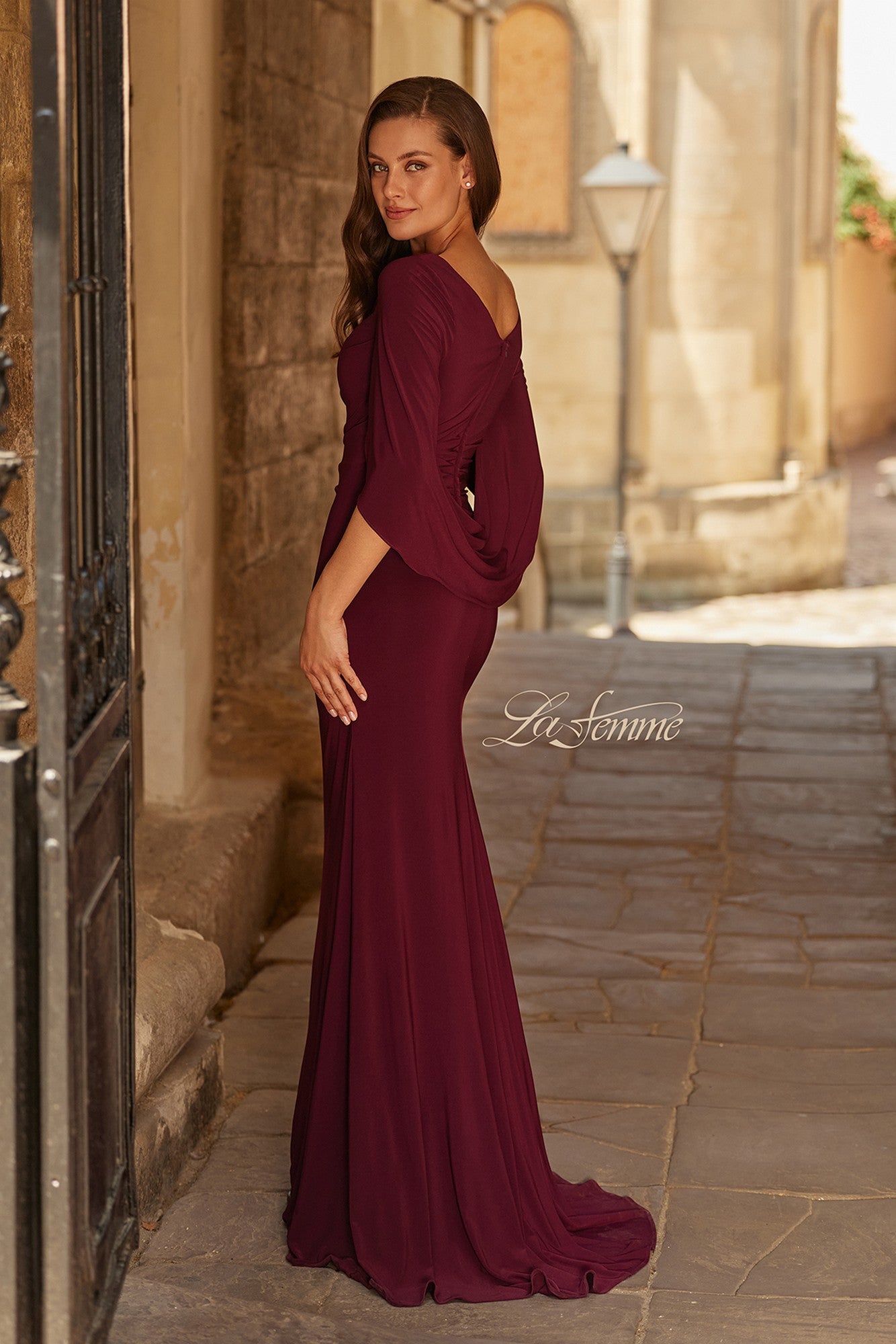 Wearing the Long Formal Dress: La Femme 33148, a woman in a burgundy jersey gown with cape-style sleeves stands on a stone pathway by a wall, glancing over her shoulder.